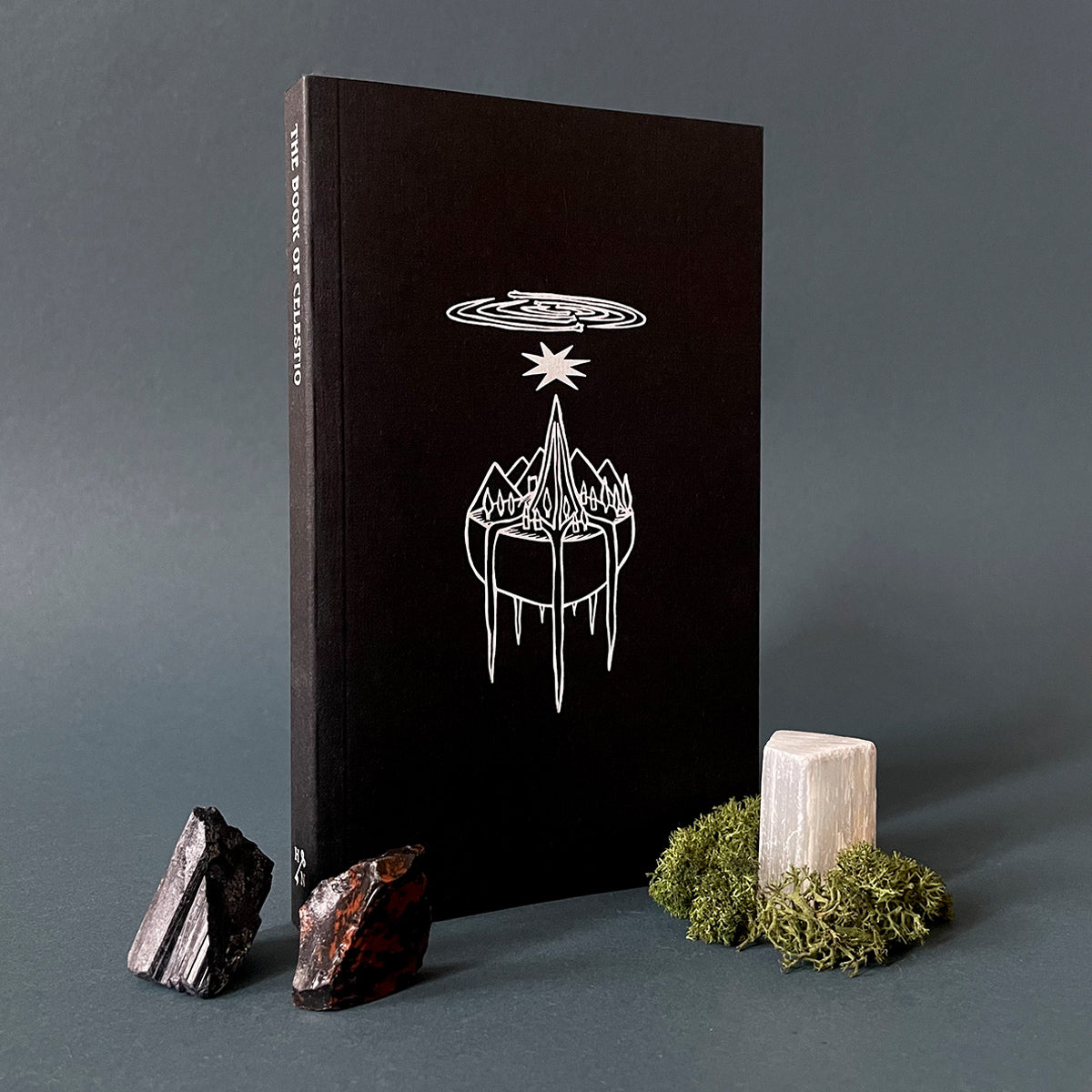 A black book with white text and an embossed design depicting a star-like figure above a planet with mountains. The book is presented upright with two small minerals and a piece of moss arranged in front.