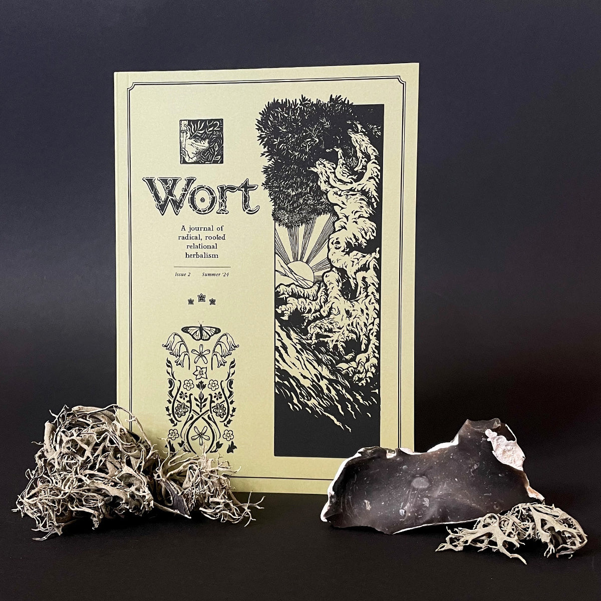 A photo of 'Wort Journal' issue 2 with a graphic design featuring plants and a figure, accompanied by a dried plant and a piece of natural-looking material, possibly part of the journal's aesthetic or content.